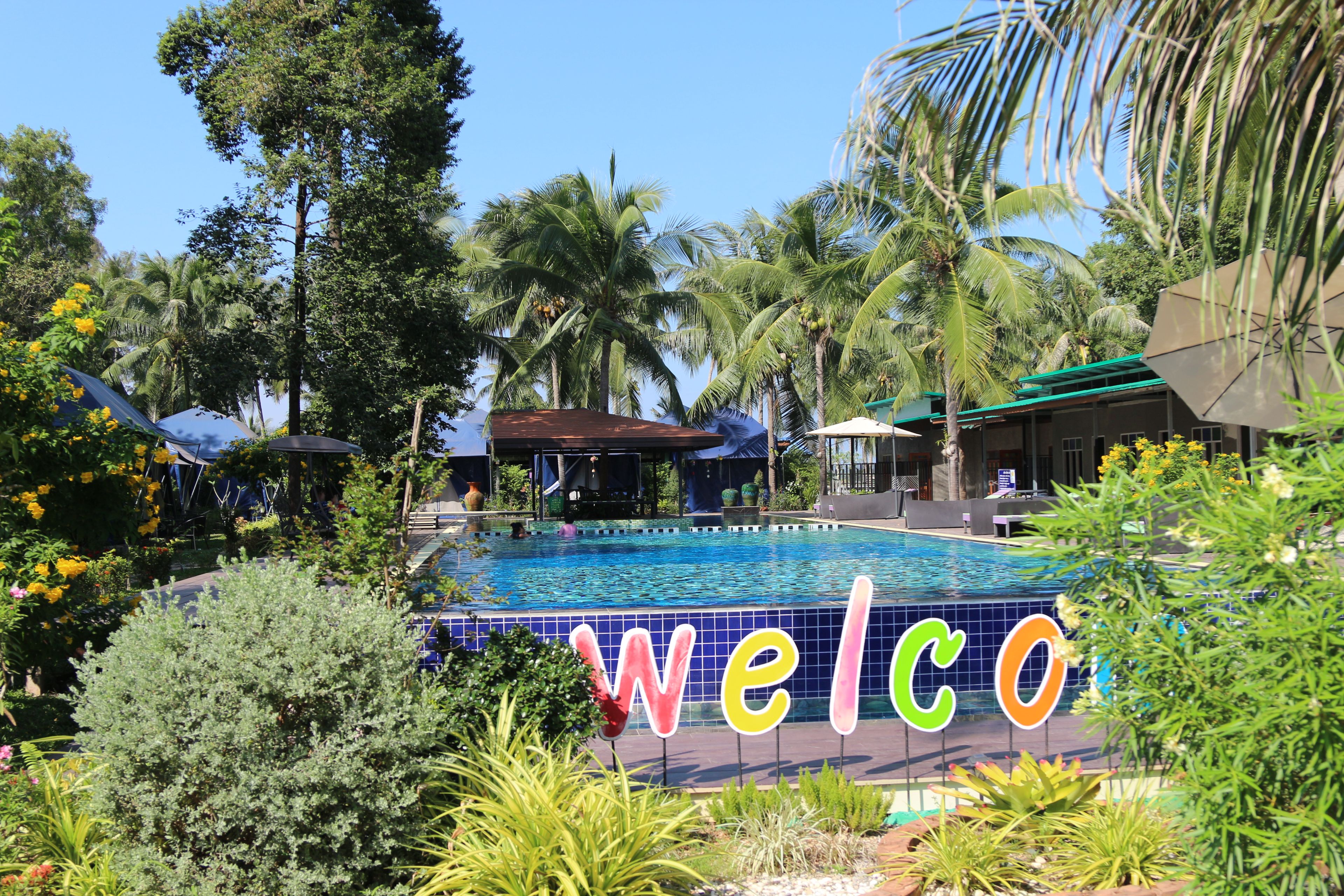 Piscina com um letreiro de 'welcome', cercada por palmeiras e plantas tropicais.
