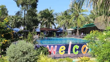 Piscina com um letreiro de 'welcome', cercada por palmeiras e plantas tropicais.