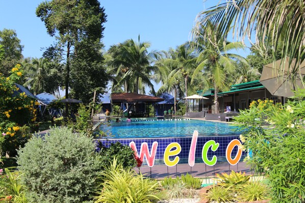 A swimming pool with a 'welcome' sign, surrounded by palm trees and tropical plants.