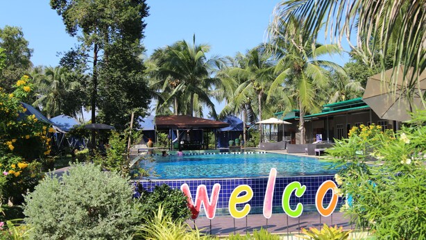 A swimming pool with a 'welcome' sign, surrounded by palm trees and tropical plants. - Moonshine Resort Chumphon (Chumphon)