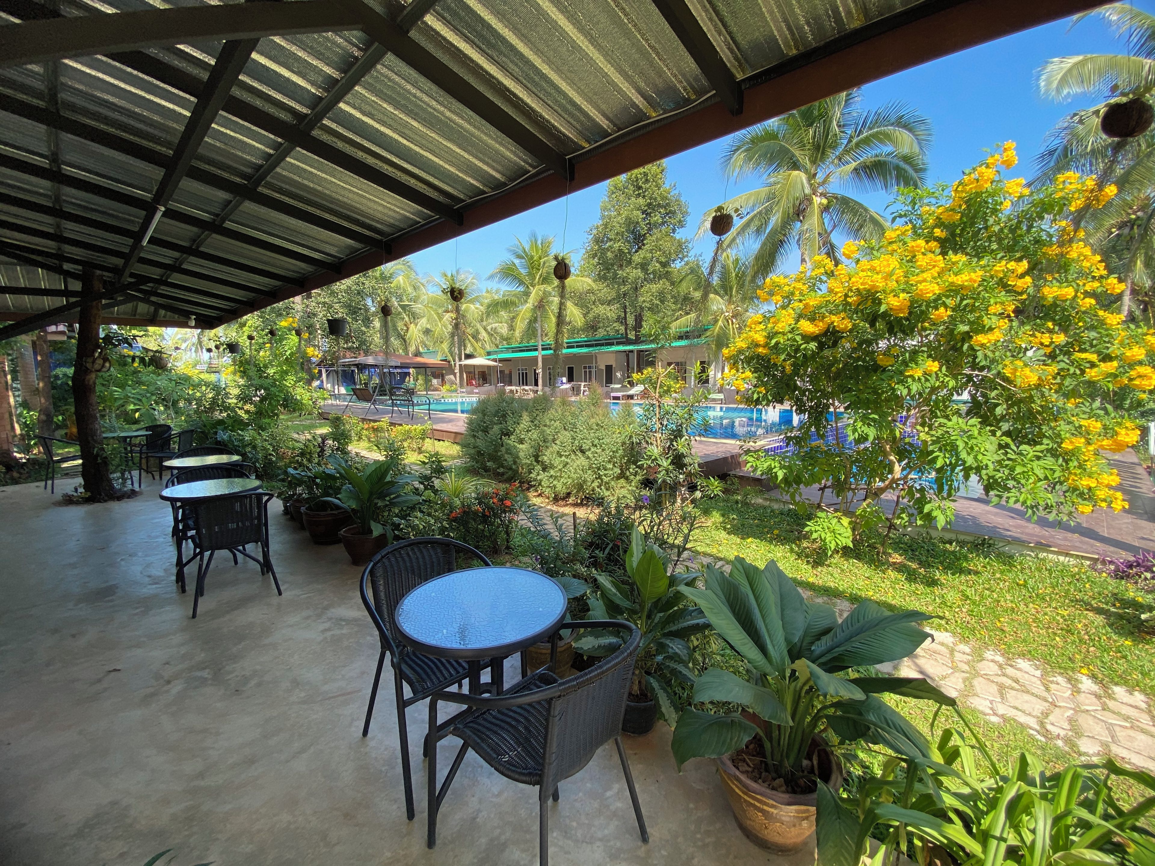 A covered outdoor area with tables and chairs, a pool, and lush greenery.