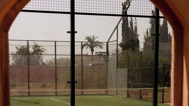 An indoor tennis court with a net, surrounded by a fence, and viewed through an arched doorway. - Jenan Mayshad (Marrakech)