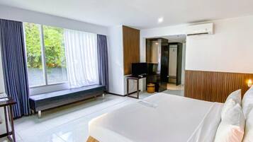 A hotel room with a large bed, a bench by the window, a desk with a chair, and an air conditioning unit.
