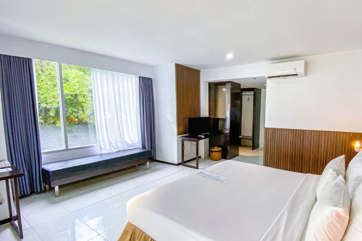 A hotel room with a large bed, a bench by the window, a desk with a chair, and an air conditioning unit.