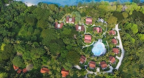 An aerial view of a resort with multiple bungalow-style buildings, a central swimming pool, and surrounding dense tropical vegetation.