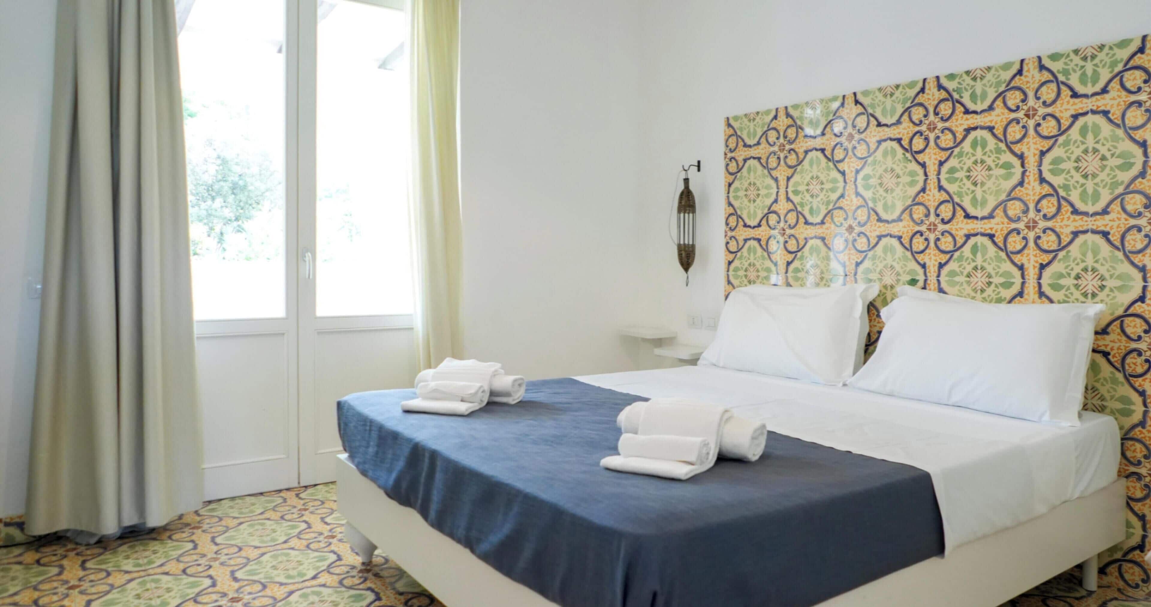 Una camera d'albergo con un letto, una finestra con tende, una parete decorata e un comodino.