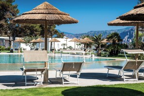 Zona de piscina con sillas reclinables y sombrillas de paja, rodeada de vegetación y con montañas al fondo.