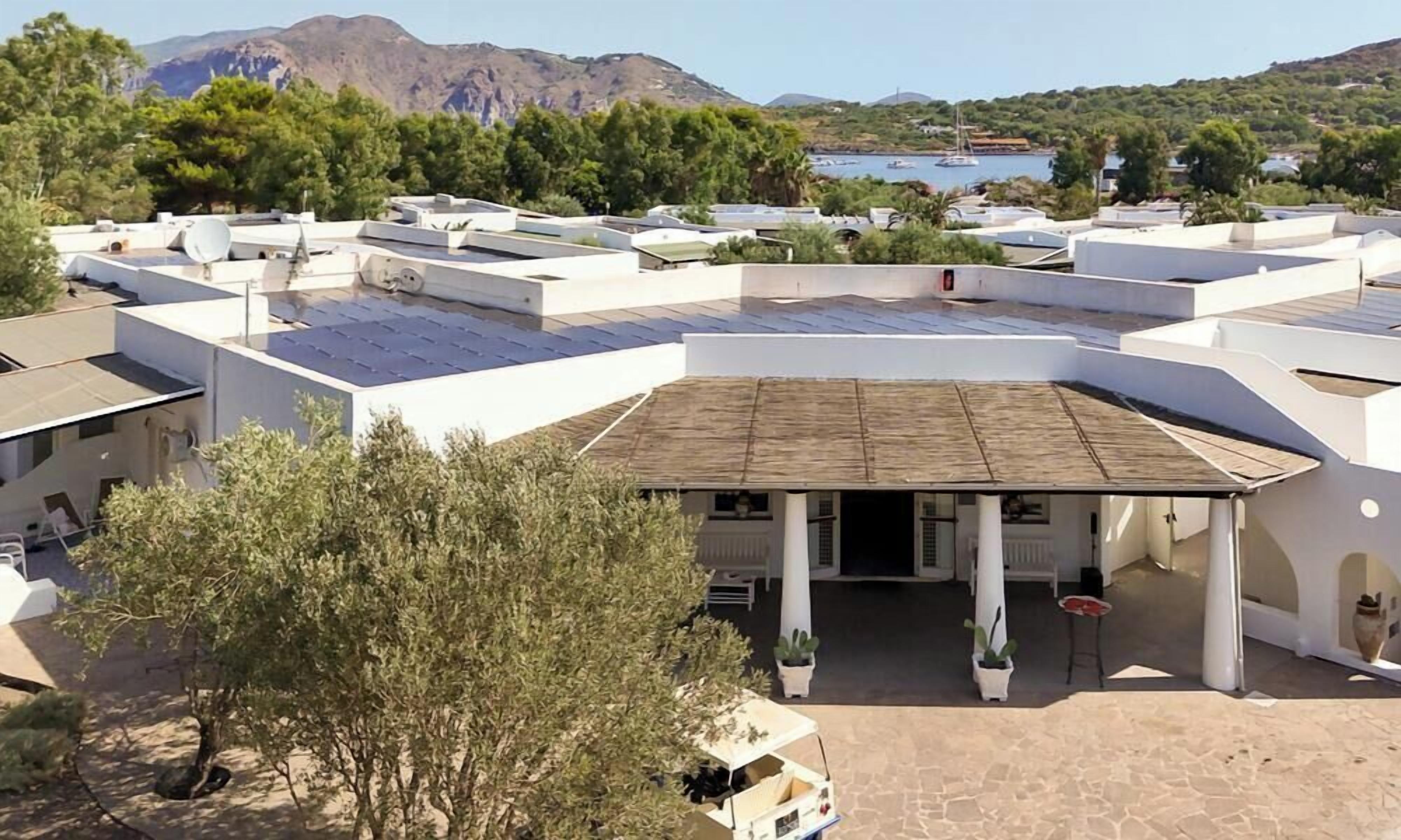 Un hôtel composé de plusieurs bâtiments aux toits blancs, doté d’un patio couvert et offrant une vue sur une étendue d’eau et des montagnes en arrière-plan.