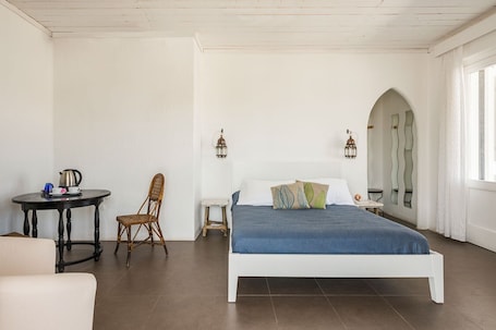 A bedroom with a bed, a chair, a small table with a kettle, and a lamp.