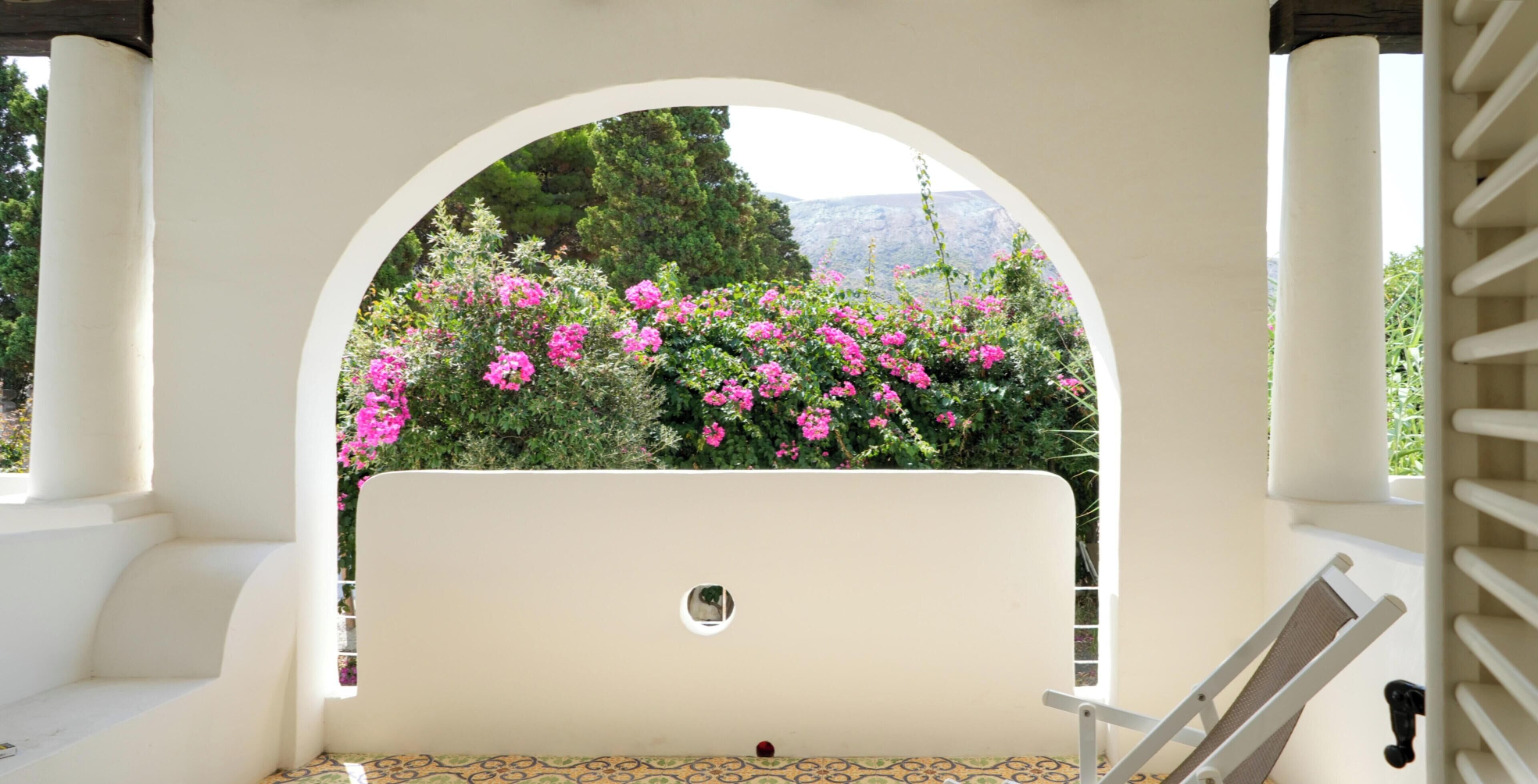 A white outdoor seating area with a bench and a table, framed by an archway with a view of pink flowers and greenery.