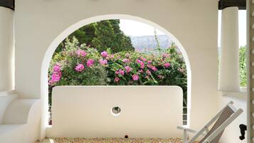 A white outdoor seating area with a bench and a table, framed by an archway with a view of pink flowers and greenery.
