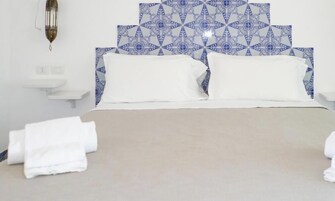 A hotel room with a bed, white pillows, a headboard with blue patterned tiles, a wall-mounted light fixture, and a white bedside table.