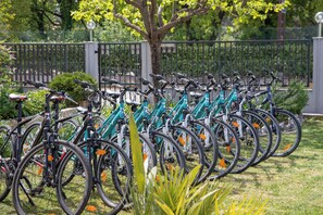 A row of bicycles parked outdoors, with a fence and greenery in the background.