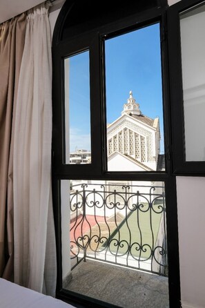 Ein Zimmer mit großem Fenster, einem Balkon mit schmiedeeisernem Geländer und Blick auf ein Gebäude mit markanter Architektur.