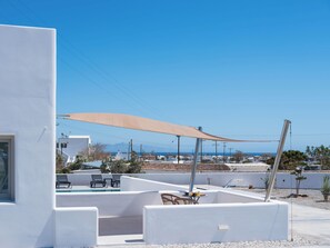 A modern outdoor pool area with a sunshade, lounge chairs, and a view of the sea and distant buildings. - Arch Suites (Santorini)
