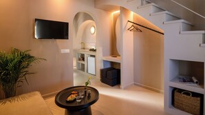 A modern interior with a flat-screen TV, a round table with snacks, a staircase, and a plant. - Arch Suites (Santorini)