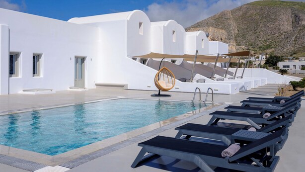 A poolside area with lounge chairs, a hanging chair, and a white building with a mountain in the background. - Arch Suites (Santorini)