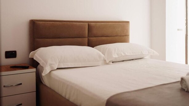 Ein ordentlich bezogenes Bett mit weißen Leinen und einem braunen Kopfteil in einem schlichten Zimmer, das einen Holfnachtschrank und eine Schubladenkommode umfasst.