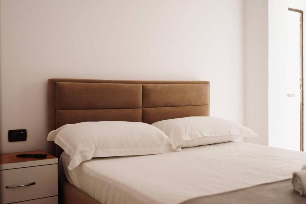 A neatly made bed with white linens and a brown headboard in a simple room with a wooden nightstand and a drawer unit.