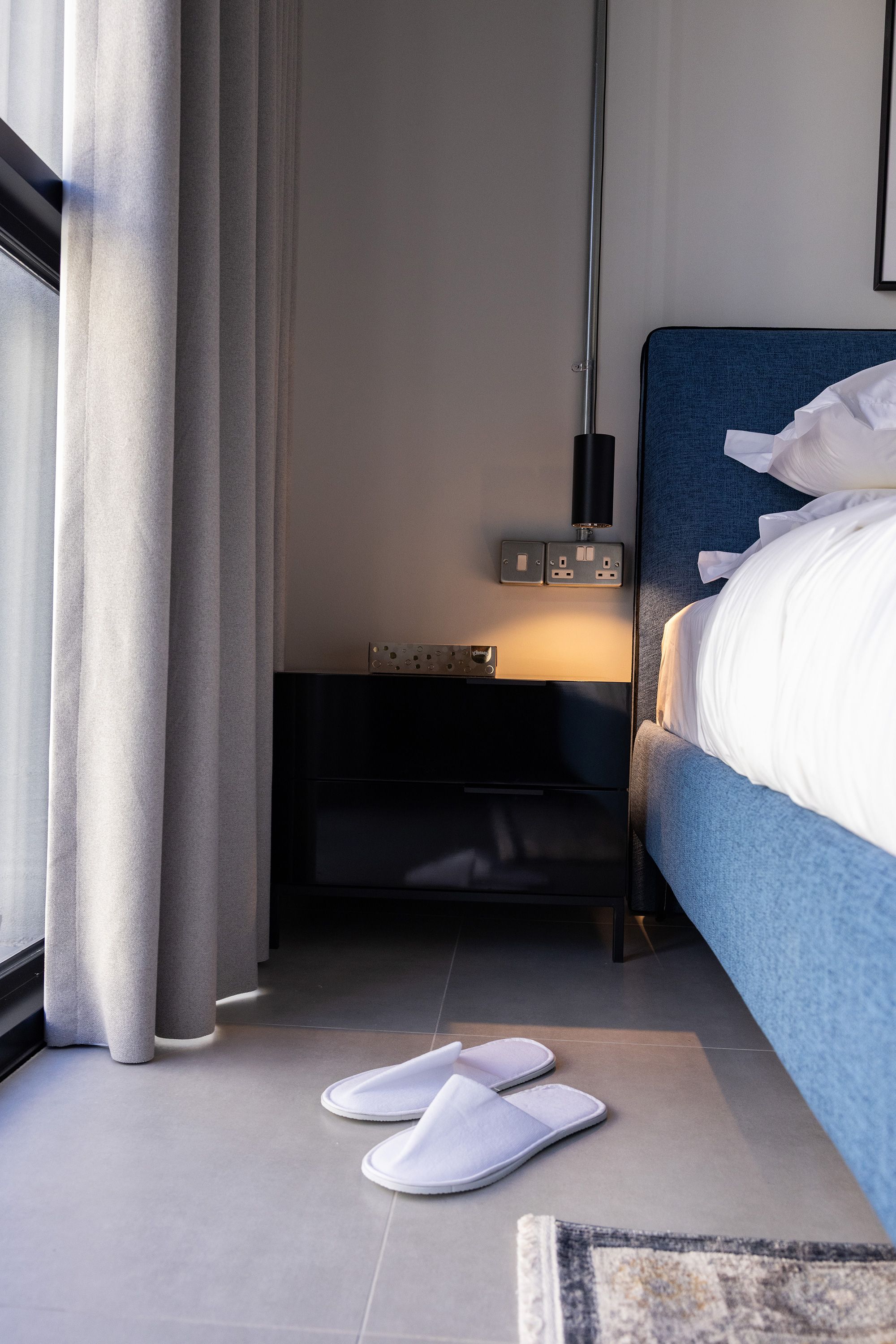 Habitación de hotel con cama, mesita de noche con lámpara, enchufe y un par de pantuflas en el suelo.