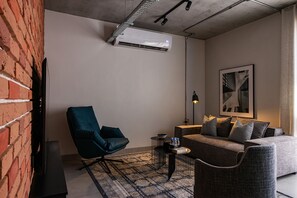 A modern living room with a brick wall, a teal chair, a gray sofa, a patterned rug, and a mounted air conditioner. - C Living 3 (Madinat Zayed)