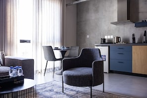 A modern living room with a sofa, a dining area with chairs, a kitchen with a stove and cabinets, and a large window with curtains. - C Living 3 (Madinat Zayed)