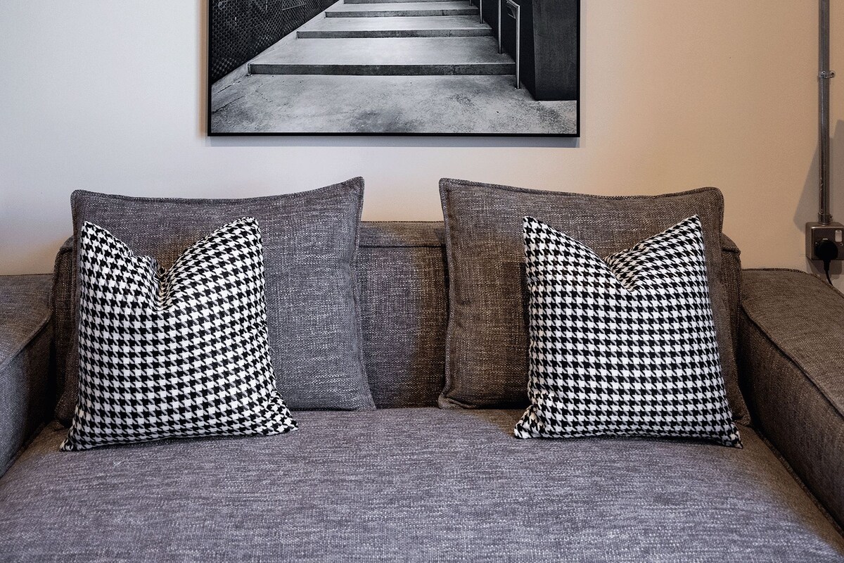 A framed artwork of a staircase on a wall, a gray sofa with patterned cushions, and a lamp on the right.