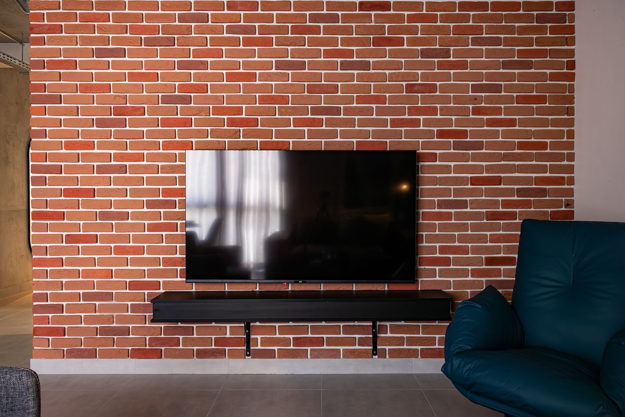 Un muro de ladrillo con un televisor de pantalla plana montado, un banco oscuro debajo y un sofá color turquesa a la derecha.