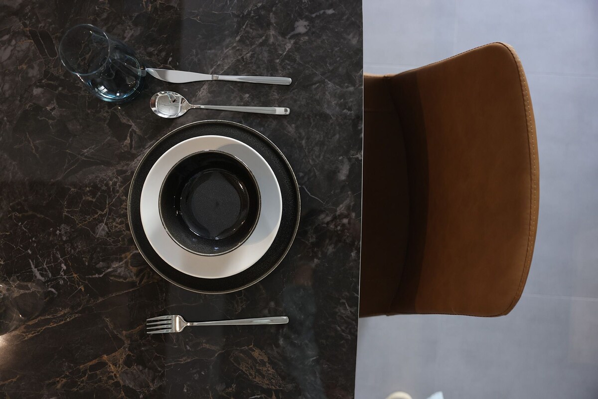 A dining table with a black and white plate, a glass, a spoon, a fork, and a knife.