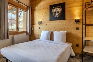 Ein Zimmer mit Holzwandverkleidung, einem Bett, einem Fenster mit Vorhängen, Wandleuchten und einem Bären-Motiv.