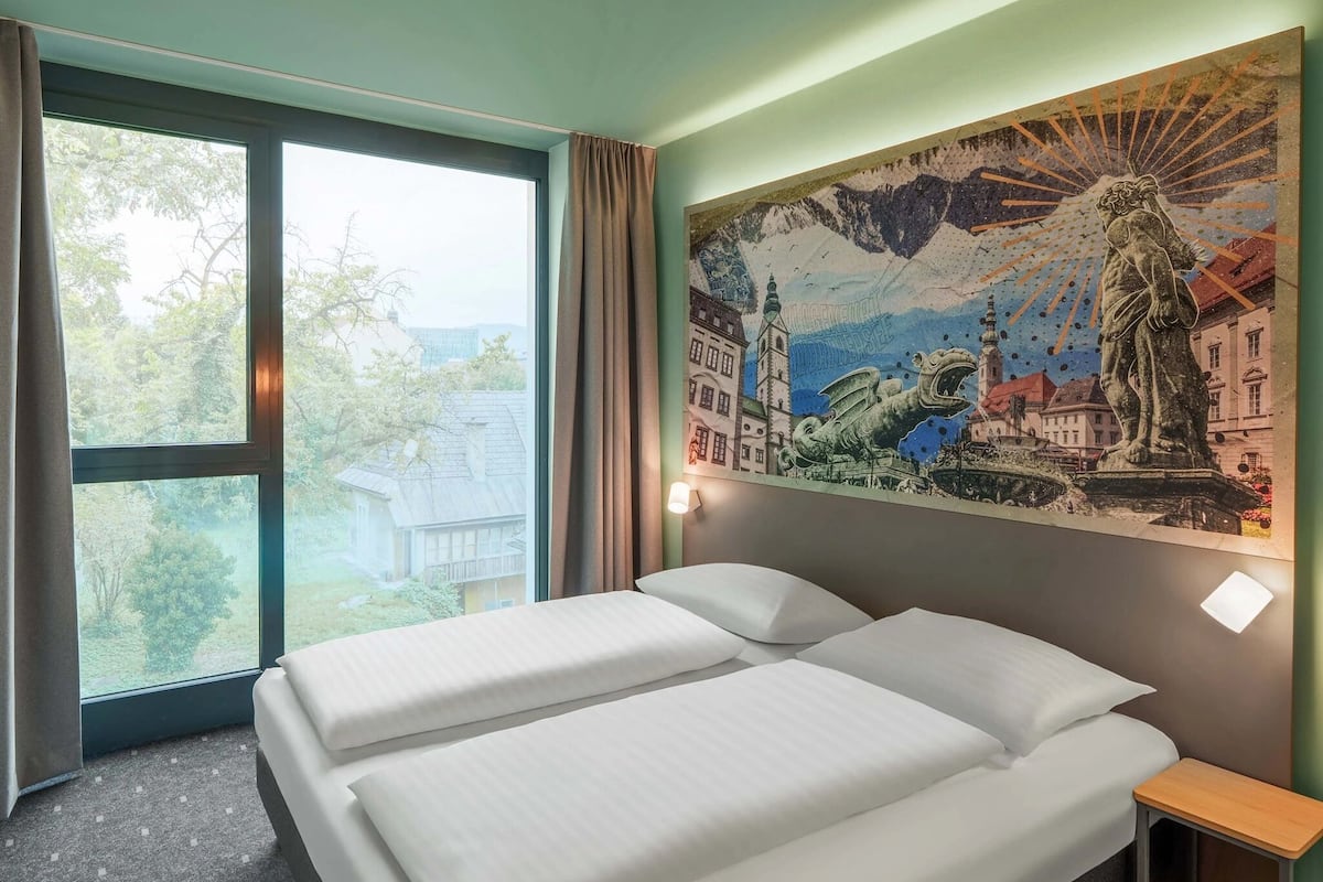 Quarto de hotel com cama, mesa de cabeceira, janela com cortinas e um mural na parede.