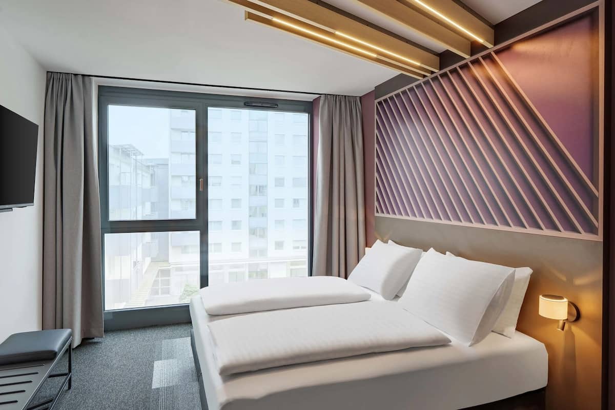 Quarto de hotel com uma cama grande, uma escrivaninha, uma TV e uma janela com vista para edifícios.