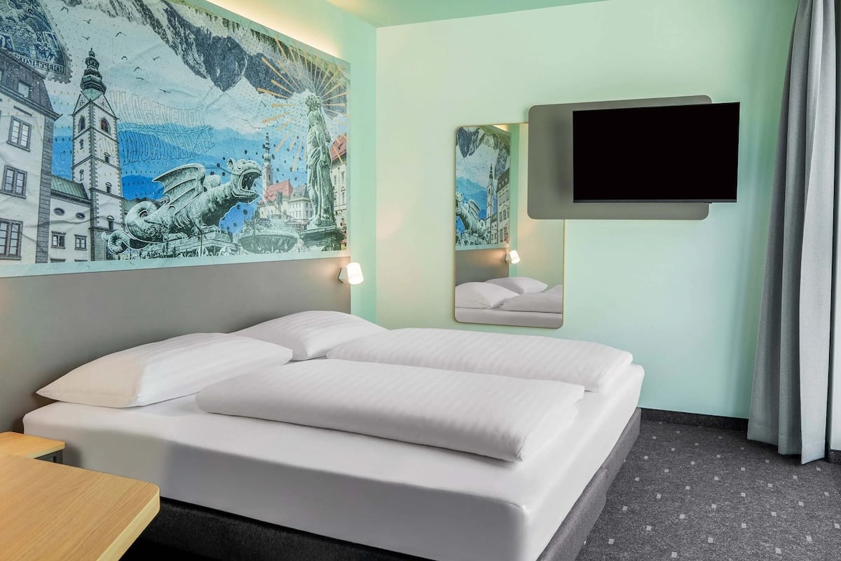 Quarto de hotel com cama, escrivaninha, televisão montada e painel na parede.