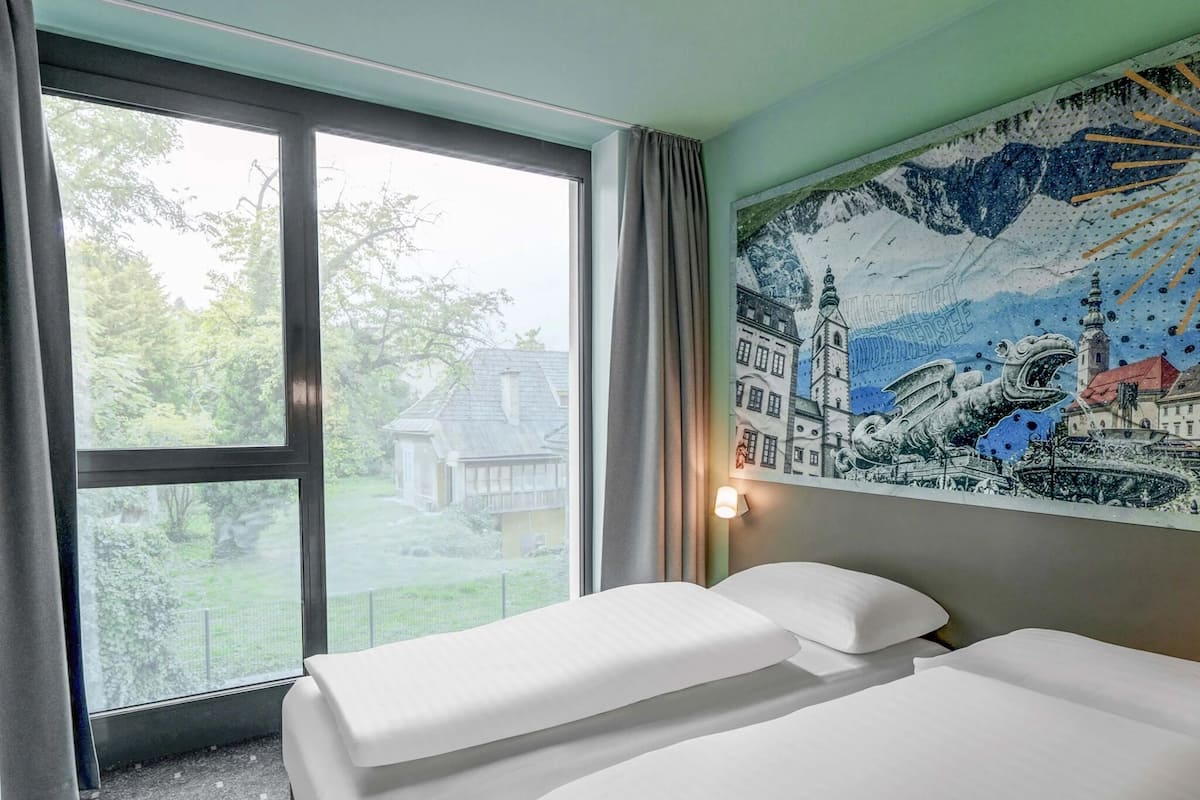 Quarto de hotel com duas camas, uma janela grande com vista para uma casa e árvores, e um mural na parede.