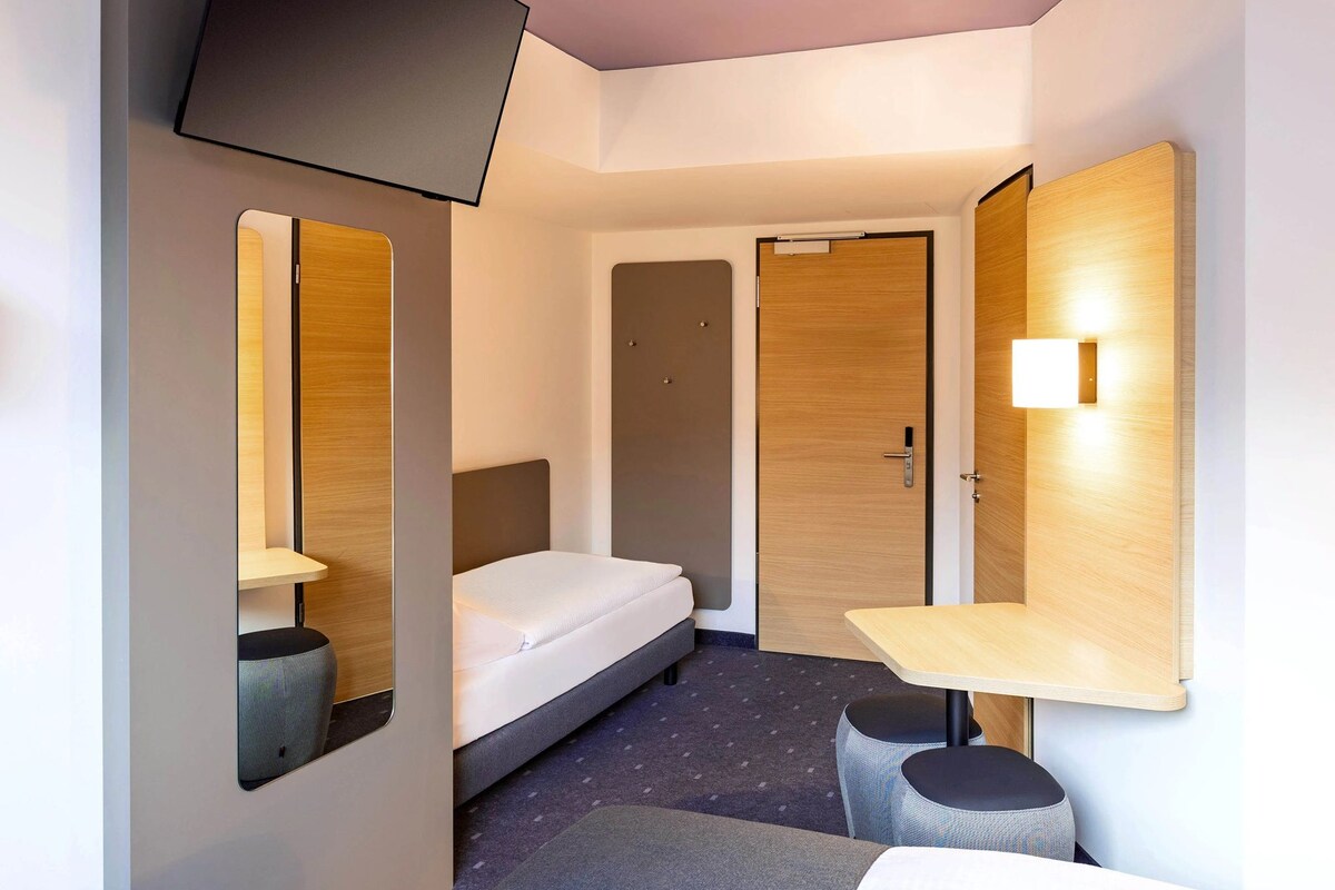 Ein kleines Hotelzimmer mit Bett, Schreibtisch, Stuhl, Spiegel und einem an der Wand befestigten Fernseher.