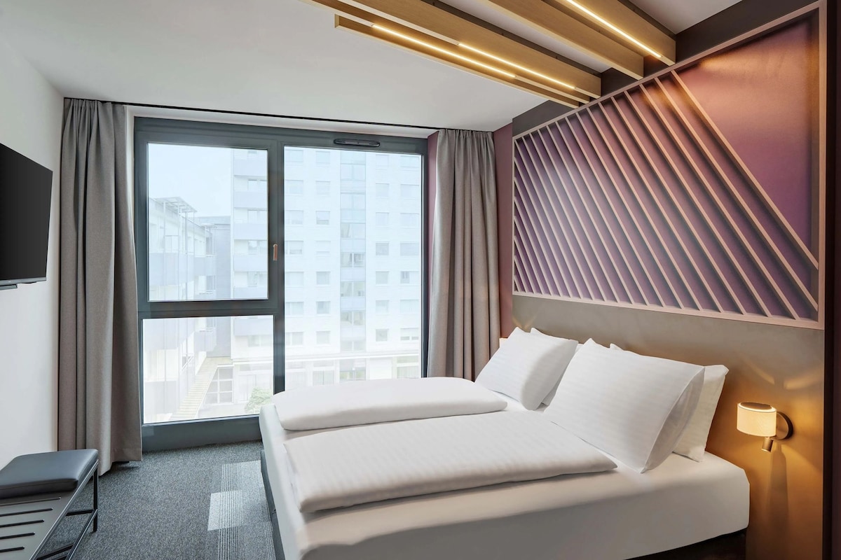 A hotel room with a large bed, a desk, a TV, and a window with a view of buildings.