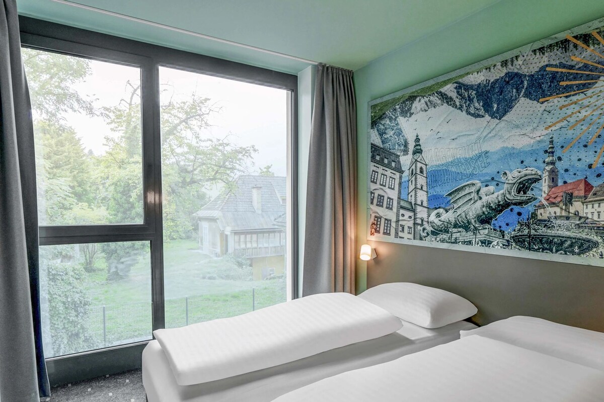 Ein Hotelzimmer mit zwei Betten, einem großen Fenster mit Blick auf ein Haus und Bäume, sowie einem Wandgemälde.