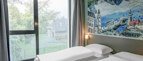 A hotel room with two beds, a large window with a view of a house and trees, and a mural on the wall.