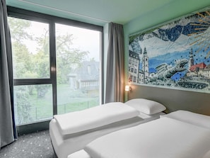 A hotel room with two beds, a large window with a view of a house and trees, and a mural on the wall.