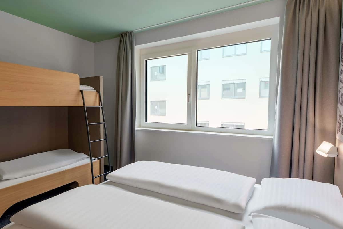 Quarto de hotel com uma cama beliche, uma janela com cortinas e vista para edifícios.