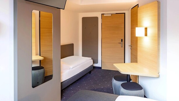 A compact hotel room with a bed, a small desk, a chair, a mirror, and a television mounted on the wall. - B&B Hotel Klagenfurt-city (Sankt Veit an der Glan)