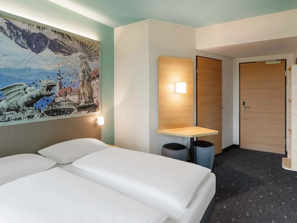 A hotel room with a bed, a wall-mounted table, a door, and a trash bin.