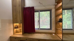 Ein Zimmer mit einem Holztisch, einem Fenster mit Blick auf Grünflächen, einem roten Vorhang und einem Regal mit einer Uhr.