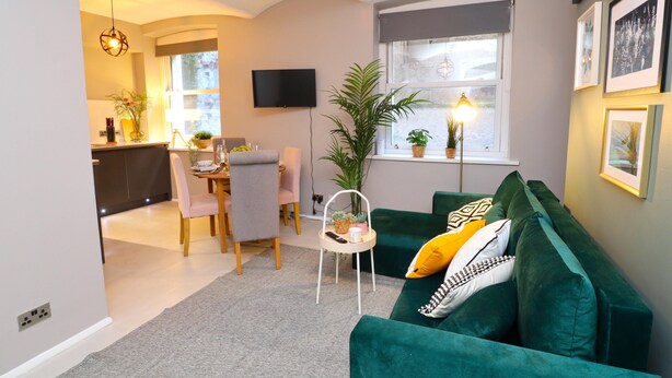 En moderne stue med en grøn sofa, et spiseområde med bord og stole, et fjernsyn ophængt på væggen, og et stort vindue med planter.