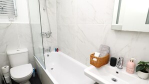 Et badeværelse med toilet, bruseniche i glas, et hvidt badekar og en vask med toiletartikler.