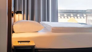 Hotellihuone, jossa on sänky, yöpöytä, lamppu ja ikkunasta avautuva näkymä rakennuksiin.