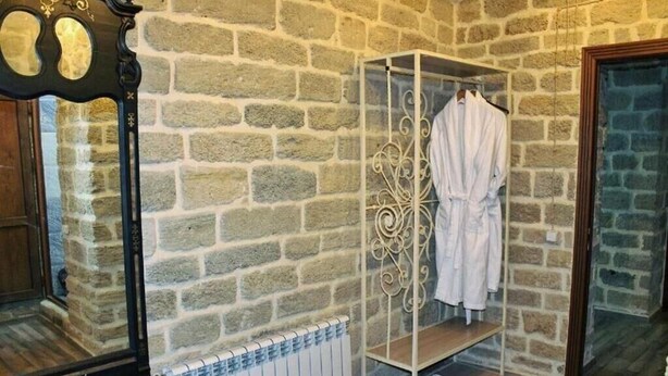 A stone-walled room with a white radiator, a wooden bench, a metal rack with a white robe, and a mirror. - Antique Palace Hotel Baku (Baku)