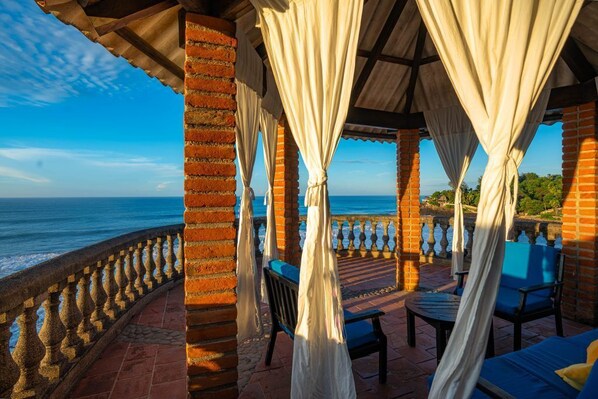 A covered outdoor seating area with brick pillars, white curtains, and a view of the ocean.