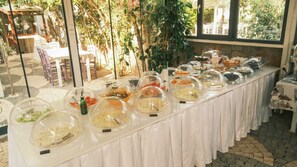 Buffet amb diversos plats coberts amb clapes, disposats sobre una taula en una sala amb grans finestres i plantes.