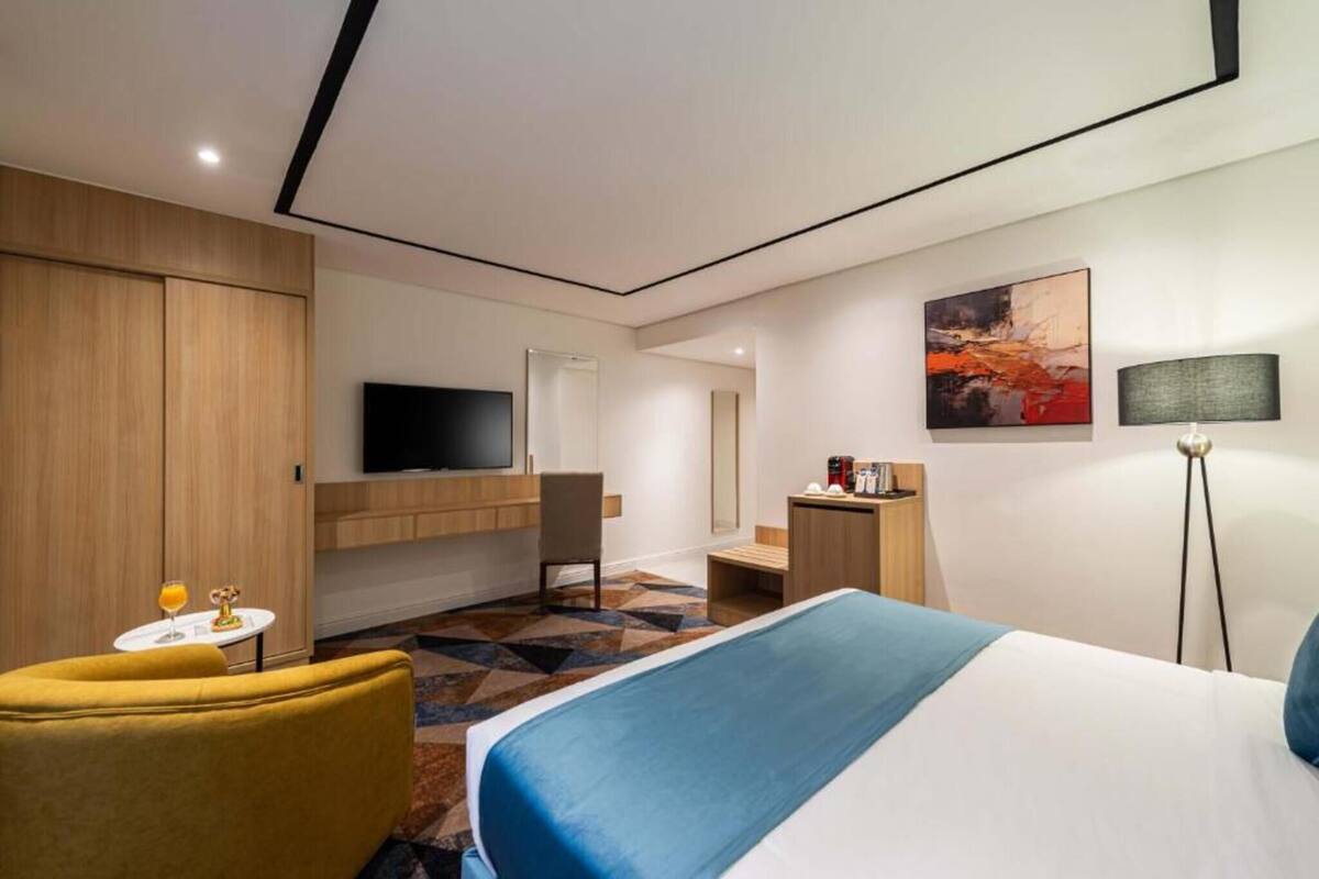 Habitación de hotel con cama, televisor, escritorio, silla y una mesita con un vaso de jugo de naranja.
